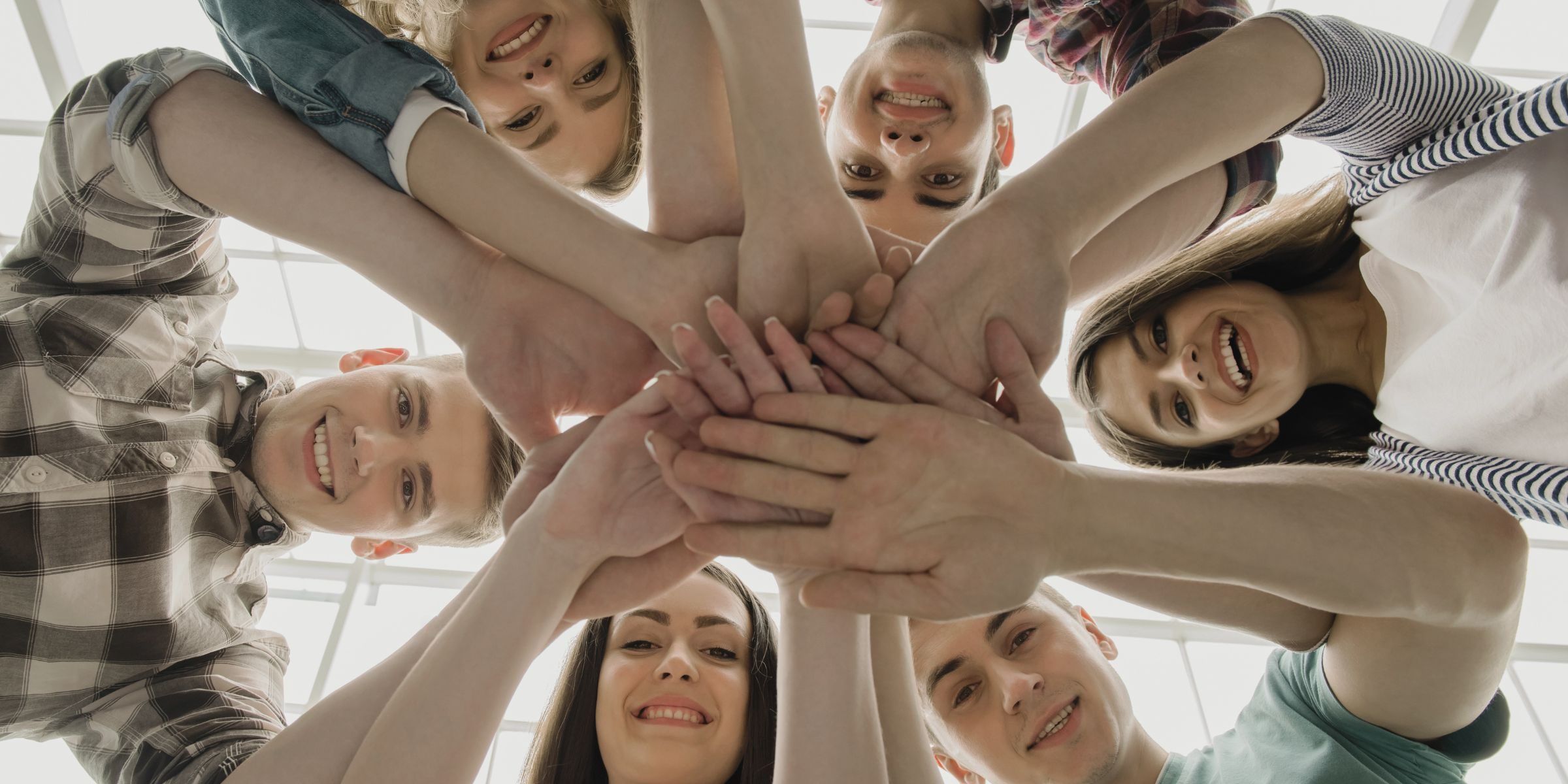 A circle of friends with hands stacked in the center, looking up at the camera, present together, eyes off their screens.